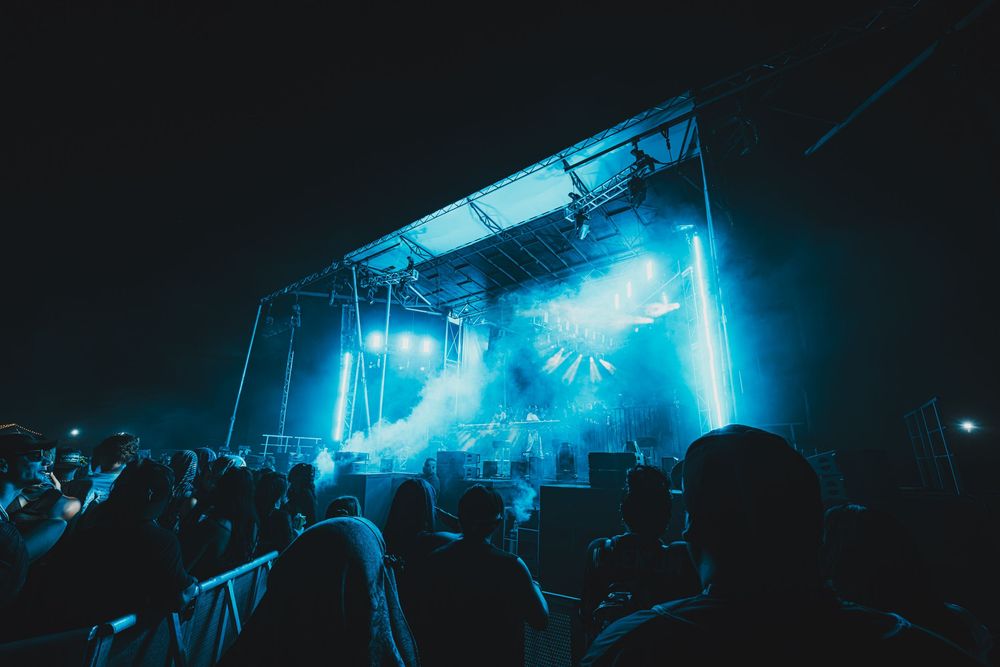 Crowd at an outdoor concert at night with bright blue stage lighting and smoke effects.
