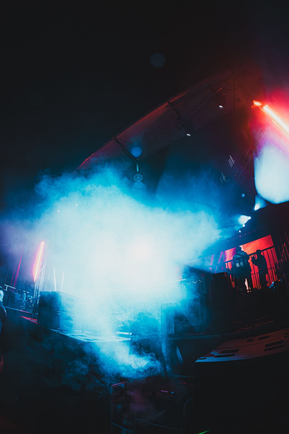Blue smoke fills a dark concert stage with colorful spotlights and silhouetted performers in a dramatic live show.