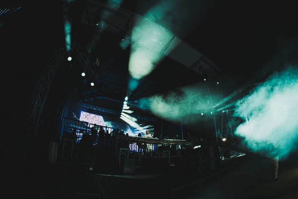 Dark concert venue with dramatic green smoke effects and stage lighting illuminating a crowd at a live music event.