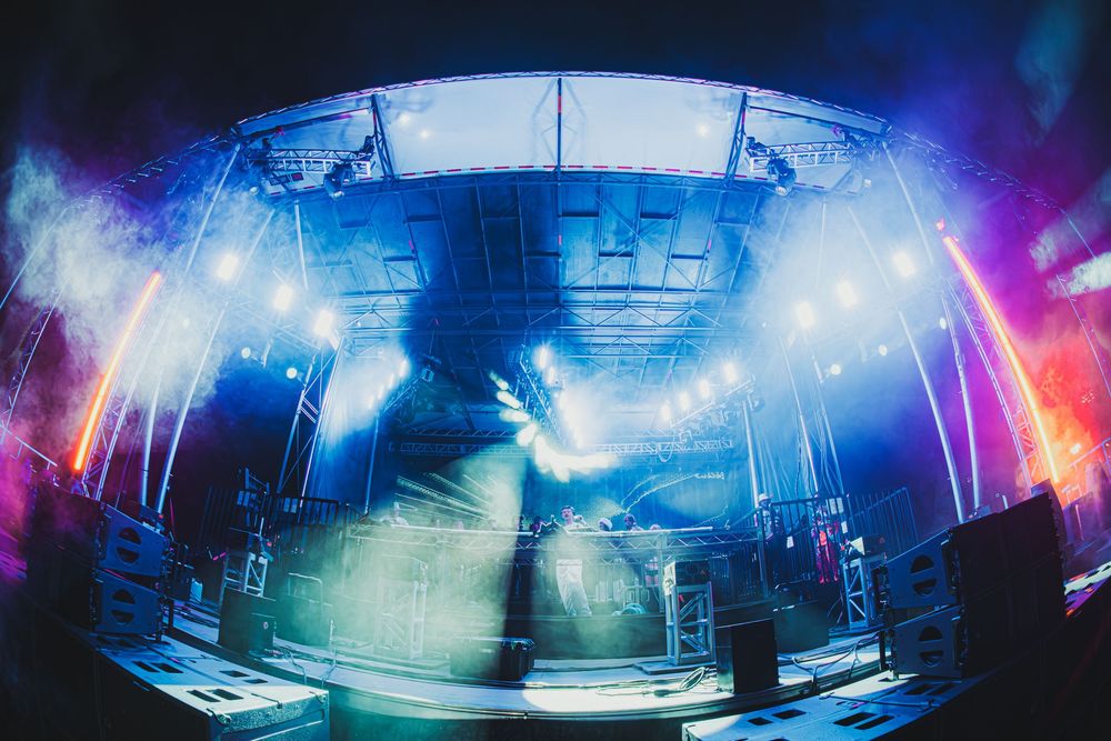 DJ performing on a vibrant stage with blue and purple lights, fog effects, and dramatic fisheye lens perspective.