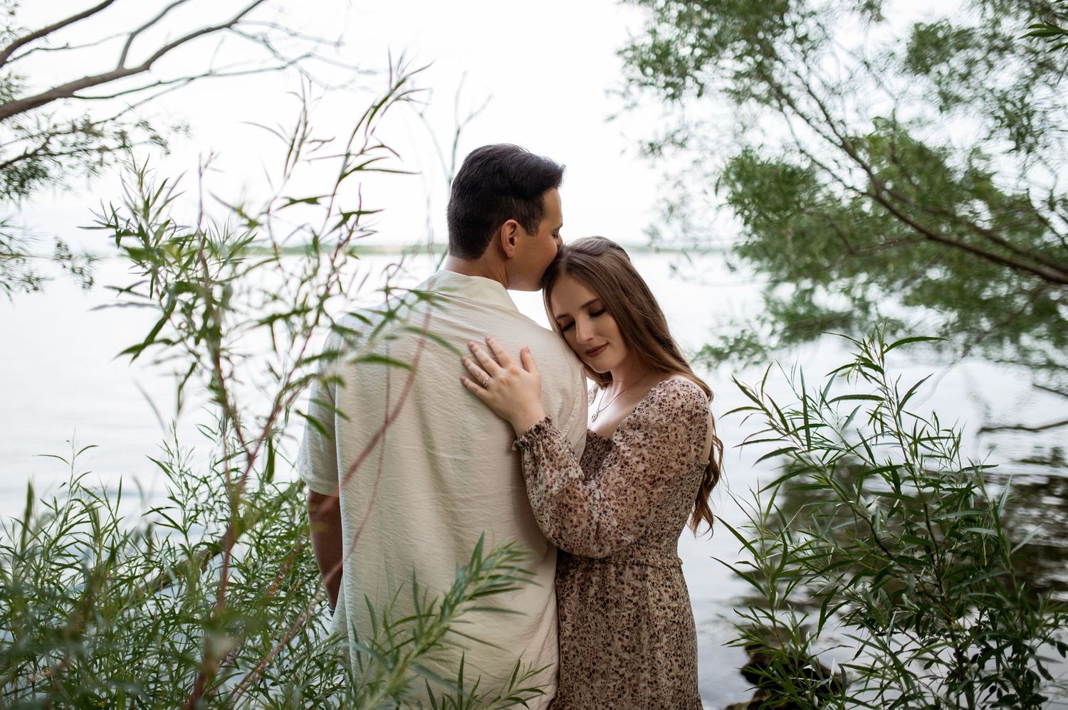 Couple embracing romantically near a serene lake surrounded by lush green trees and willow branches.