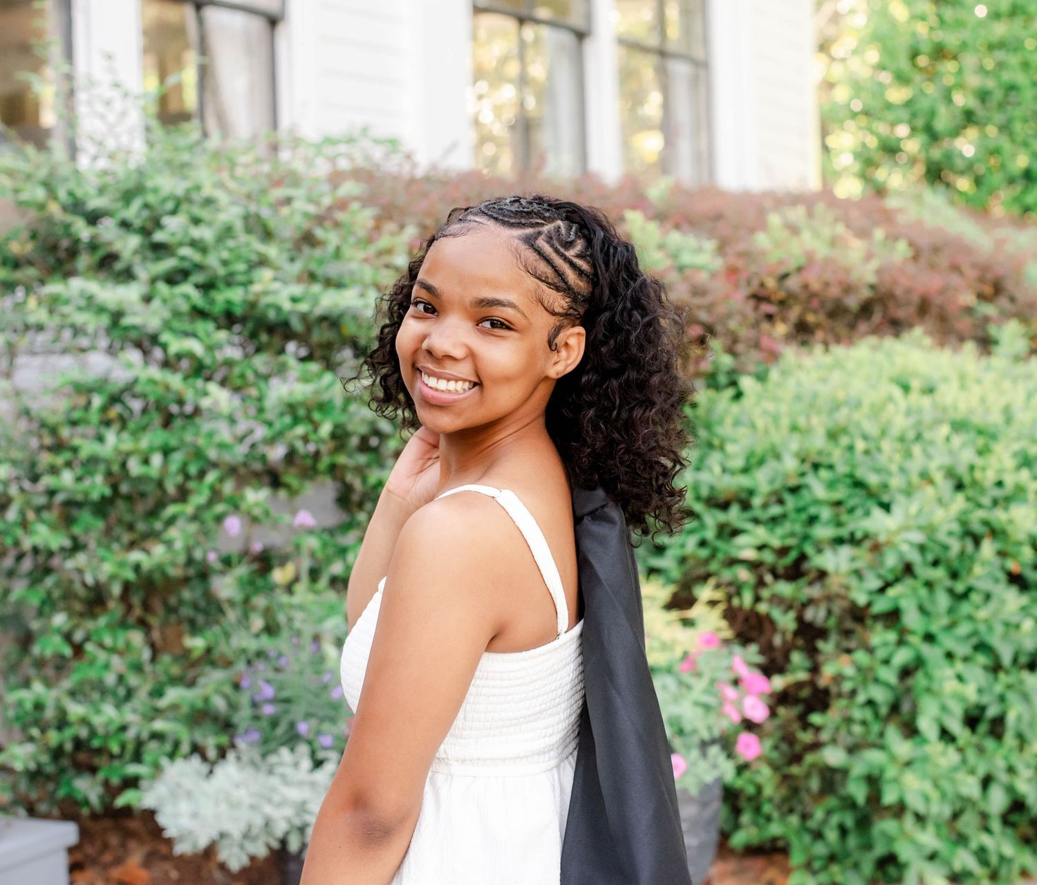 Smiling young woman in white dress holding graduation gown outdoors near green bushes and a brick building.