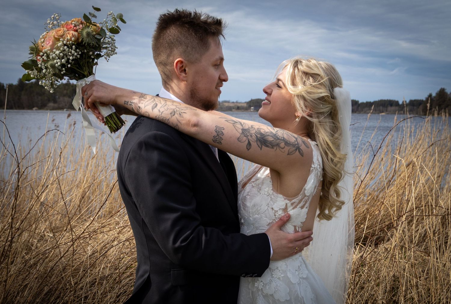 Tattooed groom embraces bride holding bouquet near waterfront in tall grass on their wedding day.
