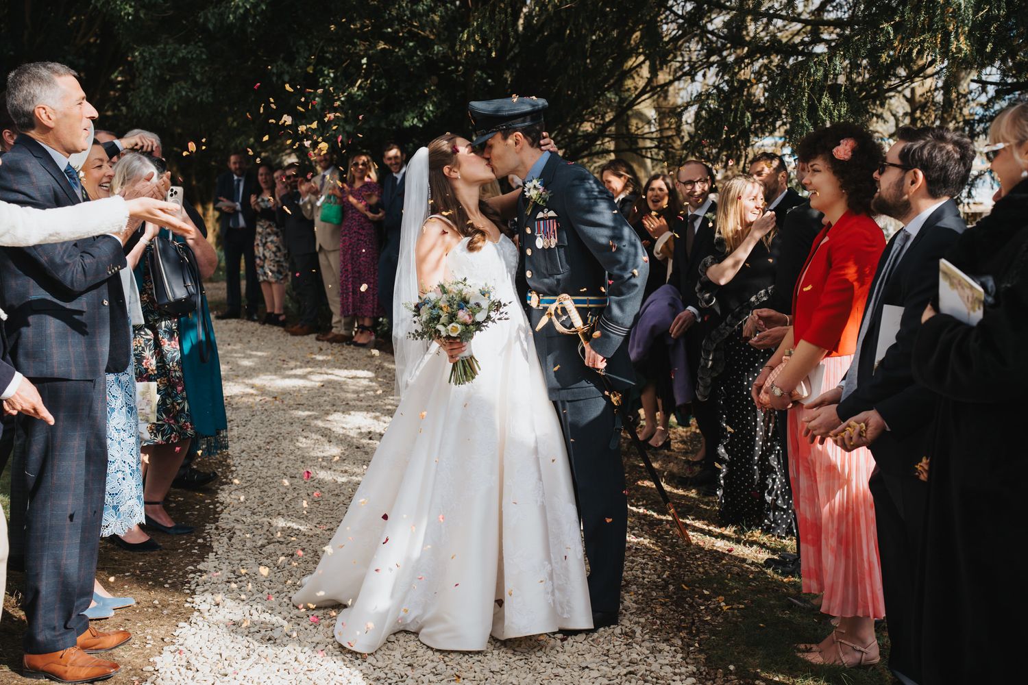 Newlyweds share first kiss as married couple while guests throw confetti along tree-lined path.