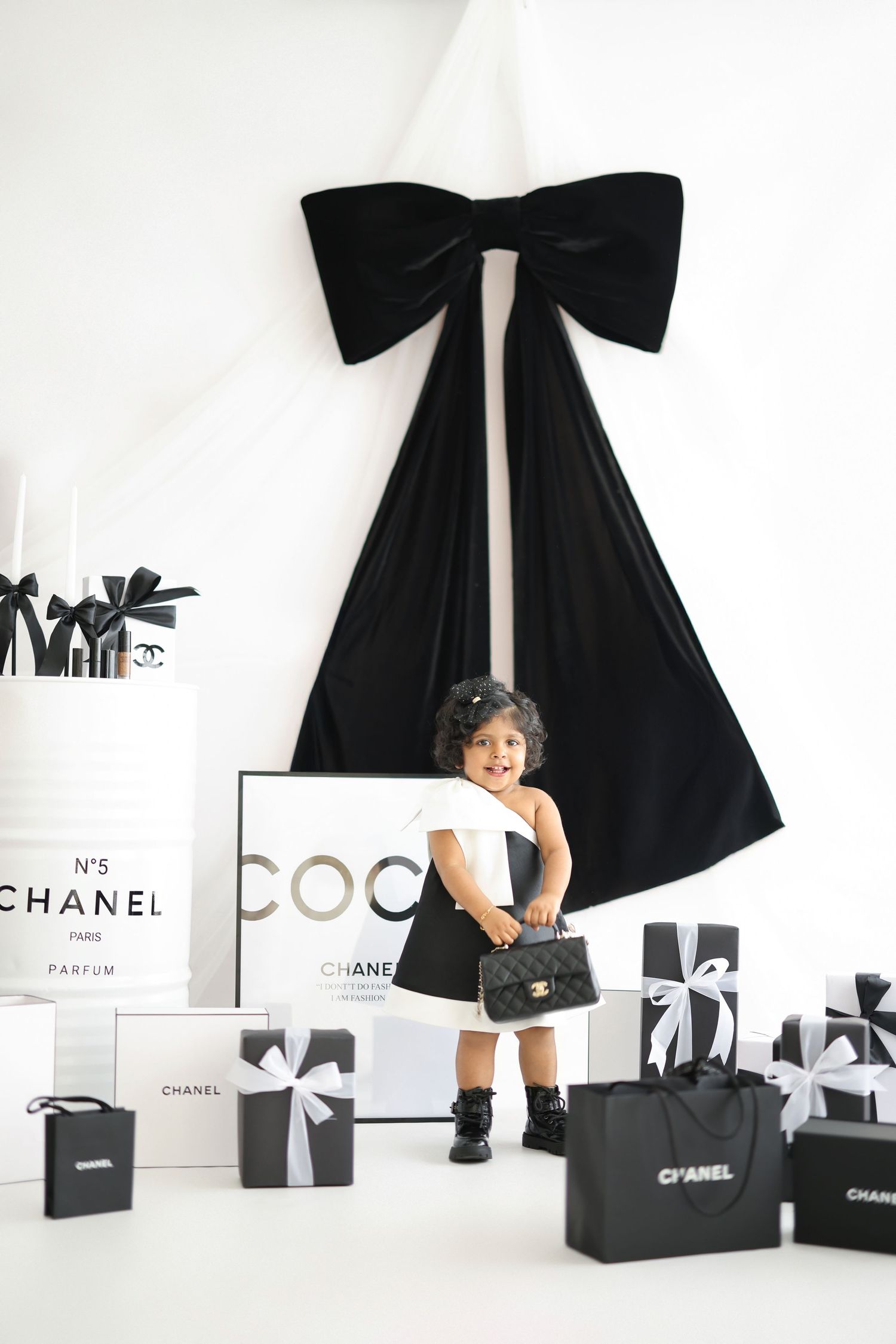 Toddler girl in black and white outfit holding Chanel bag surrounded by Chanel gift boxes and large black bow decor.