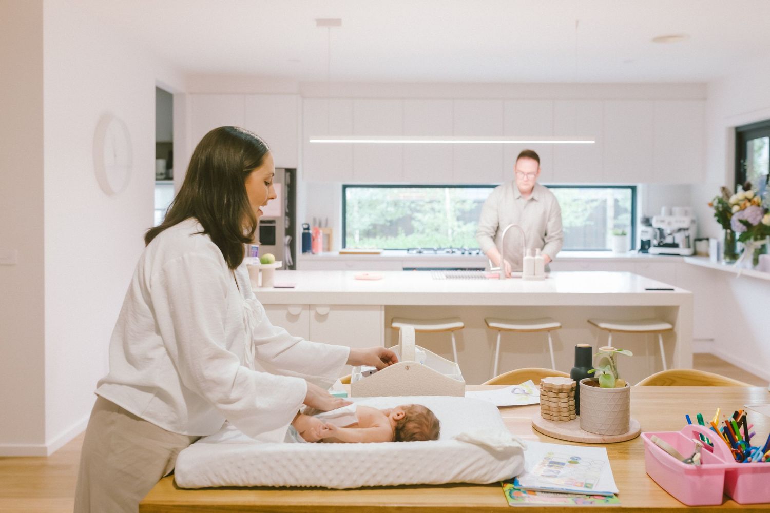 Mother changing baby on table while father stands at kitchen counter in modern white home.