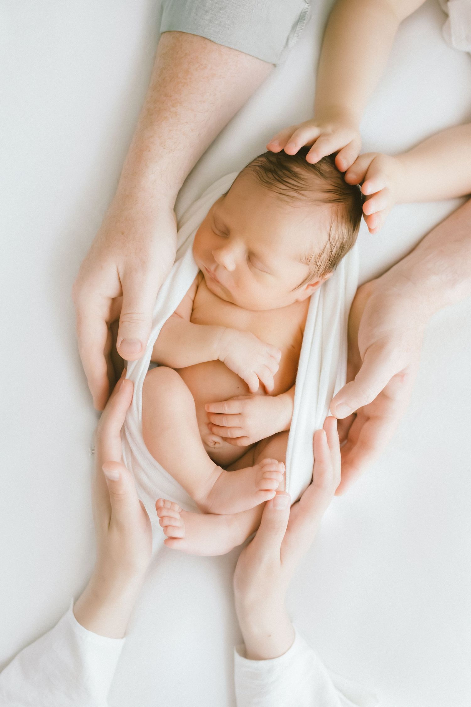 Parents cradling a sleeping newborn baby wrapped in white cloth, tender family moment captured from above.