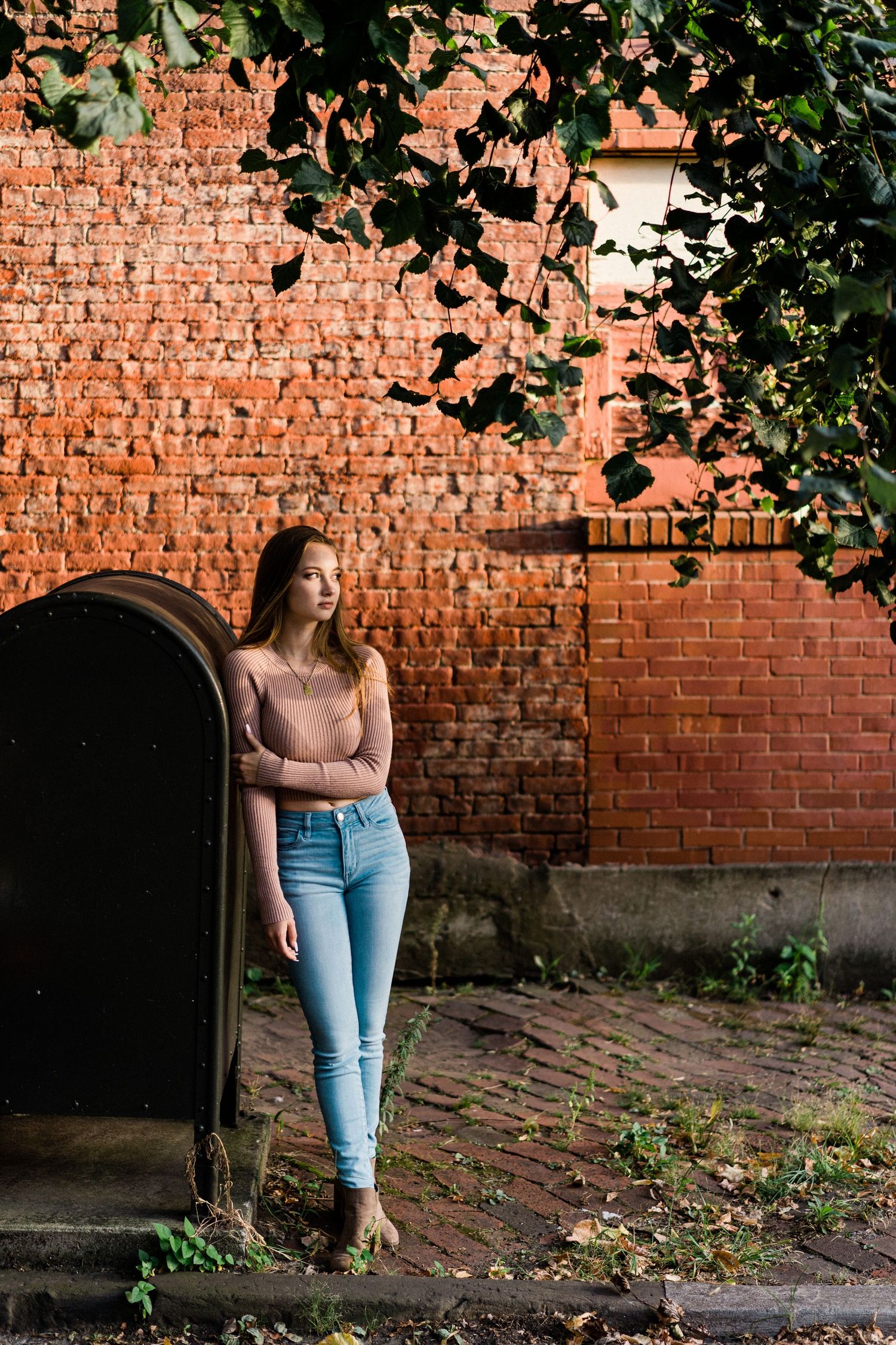 Young woman in pink top and blue jeans leaning against arched door by red brick wall with green foliage overhead.
