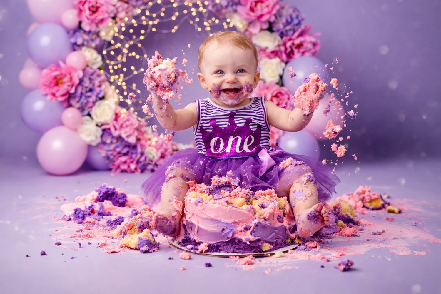 Baby girl in purple dress smashes first birthday cake surrounded by flowers and balloons in a cake smash photo session.