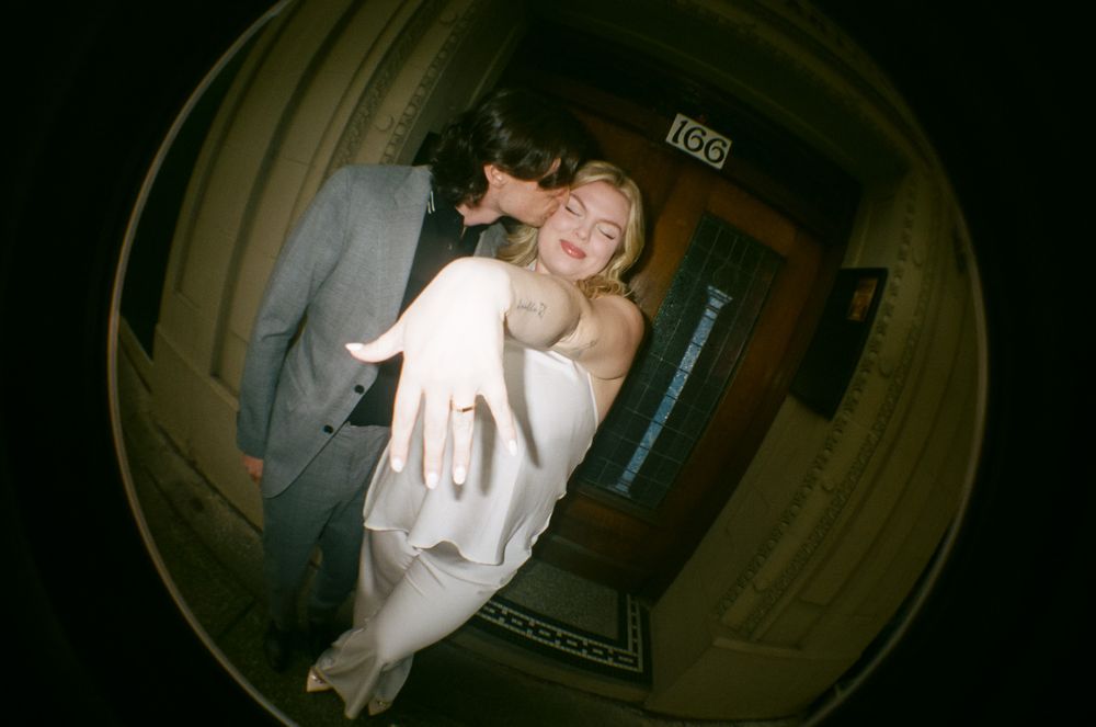 Couple kissing at doorway, woman in white dress gesturing toward camera, fisheye lens photo.