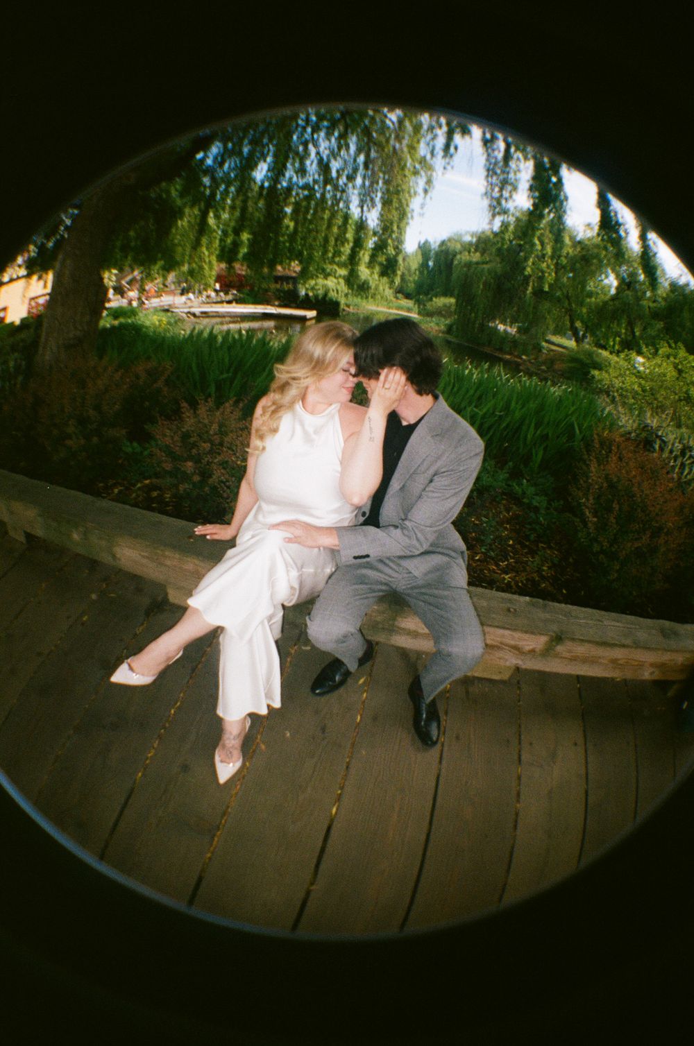 Couple sharing a romantic kiss on a wooden deck surrounded by lush green willow trees, shot with fisheye lens.