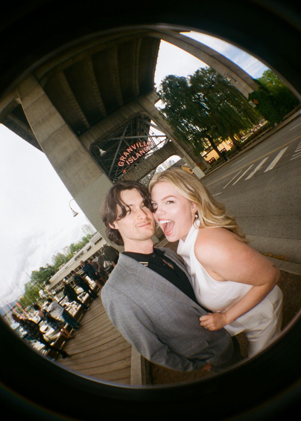 Playful couple posing under Granville Island bridge in Vancouver, laughing together in a fun fisheye photo.