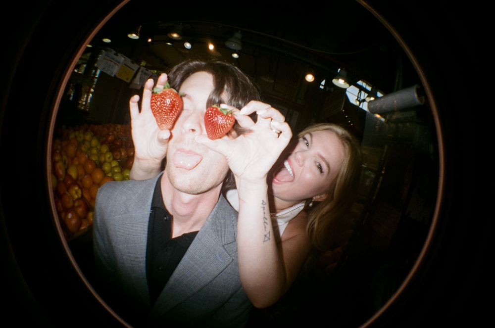 Couple playfully holding strawberries over their eyes at a colorful fruit market in a fisheye lens shot.