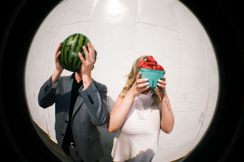 Two people humorously hiding their faces behind a watermelon and a bowl of watermelon slices indoors.