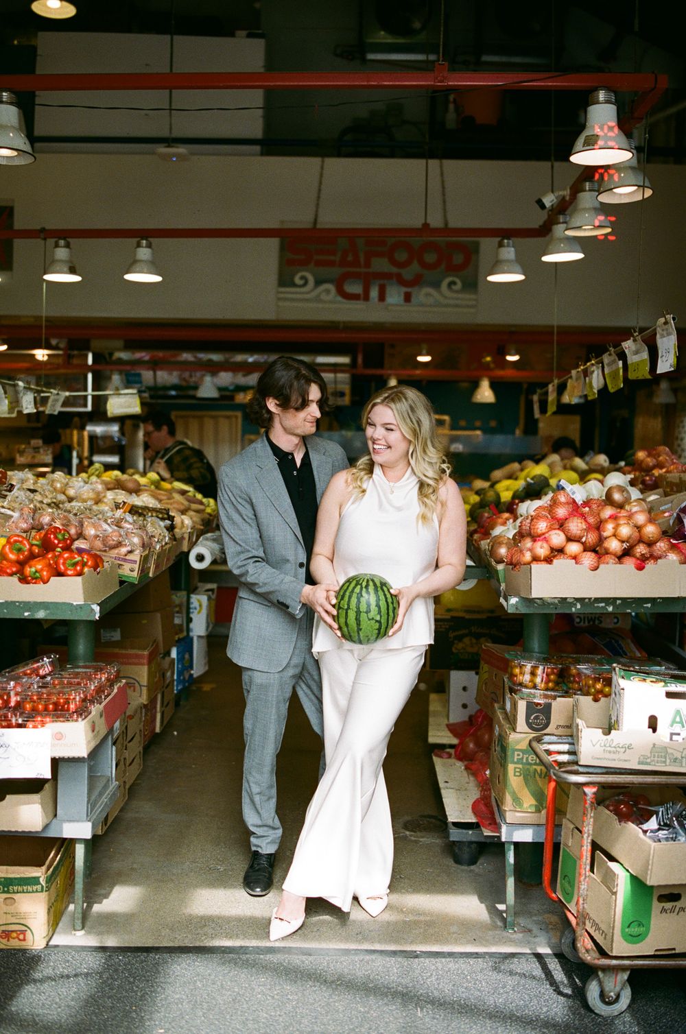 Couple holds watermelon while posing in colorful produce aisle at Seafood City grocery store engagement photo.