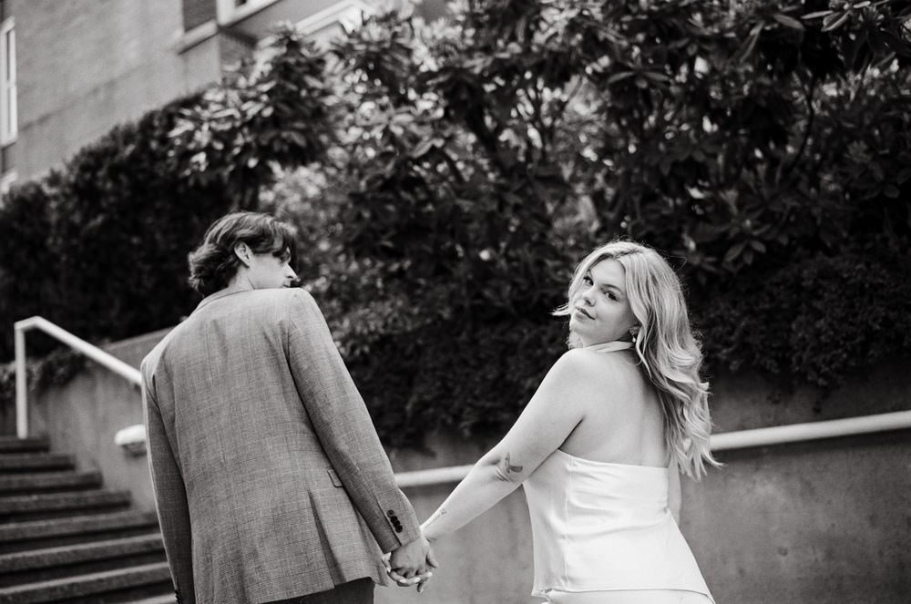 Black and white photo of a bride and groom holding hands, walking up steps, as the bride looks back smiling.