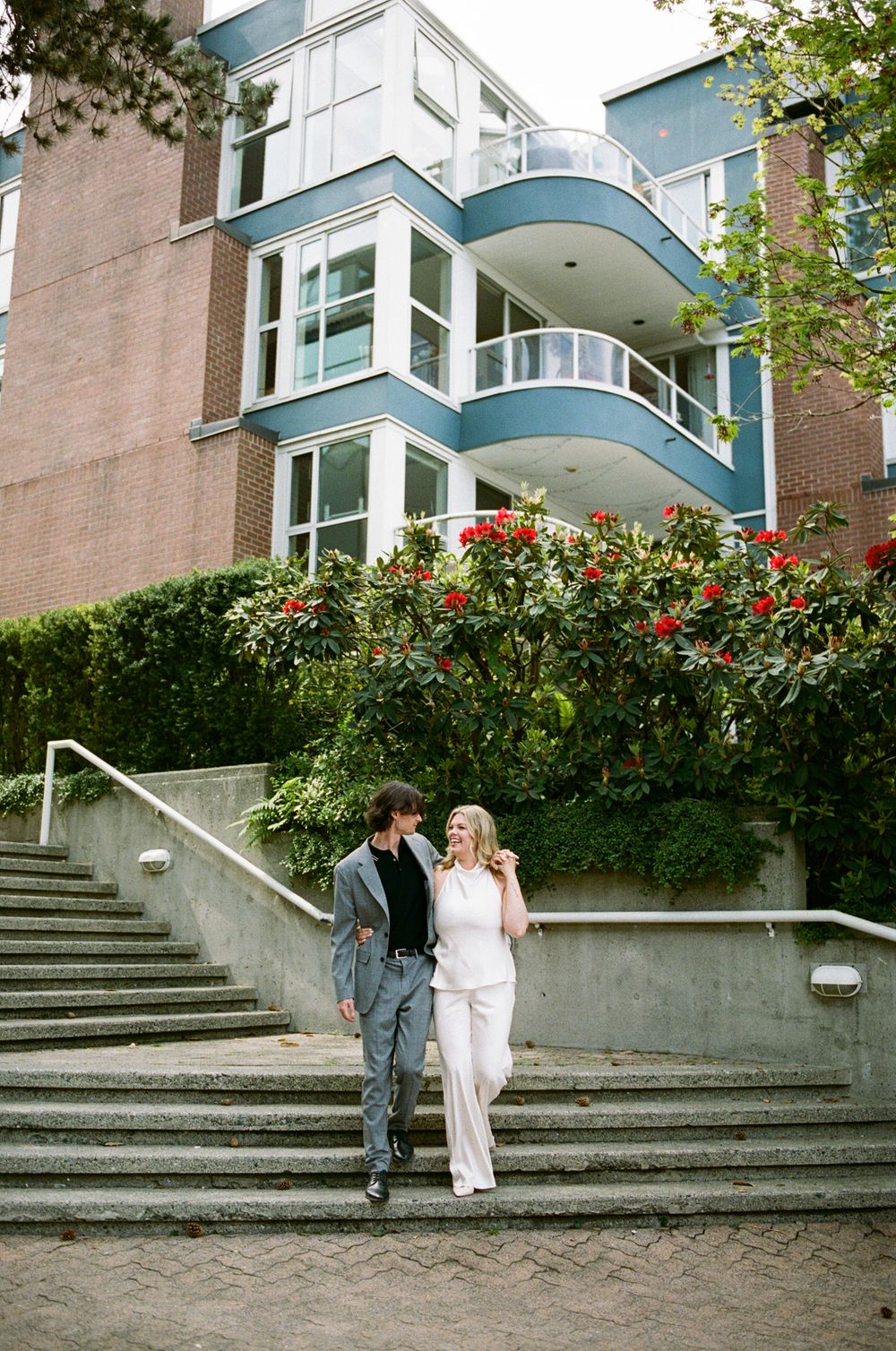 Couple in formal attire walking hand in hand in front of a modern blue building with lush greenery and red flowers.