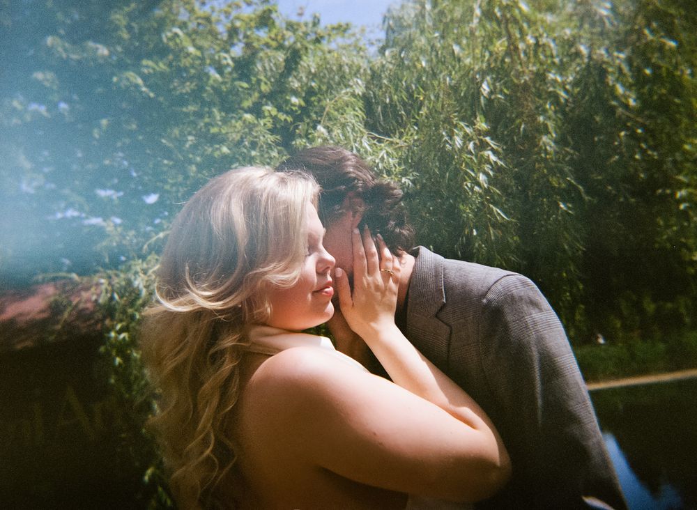 A couple shares a romantic kiss outdoors surrounded by lush green trees in soft natural light.