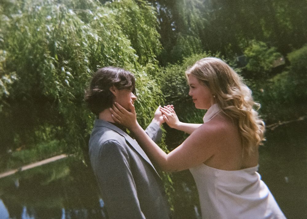 Romantic couple sharing a tender moment by a serene pond under weeping willow trees on a sunny day.