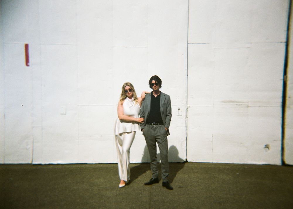 A stylish couple poses against a white wall outdoors, woman in white pantsuit and man in gray suit.
