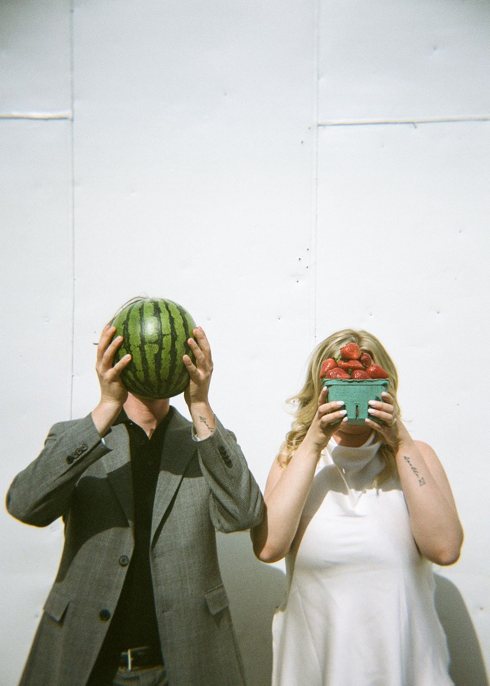 Couple playfully hiding their faces behind a watermelon and bowl of strawberries against a white wall.