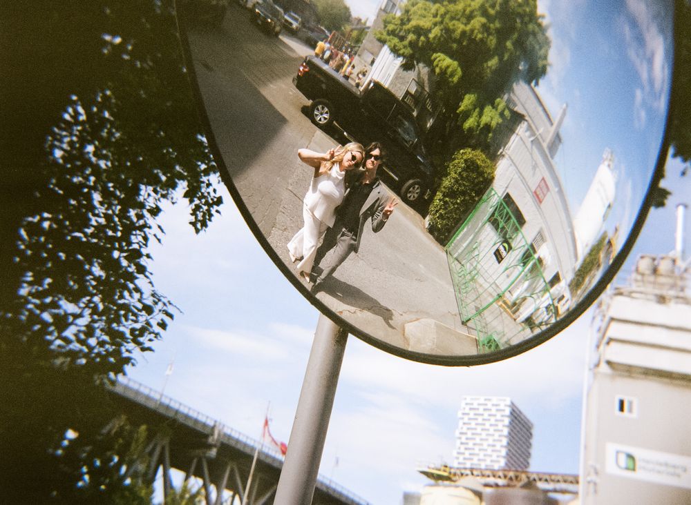 A couple reflected in a convex street mirror on a sunny urban street with trees and buildings.