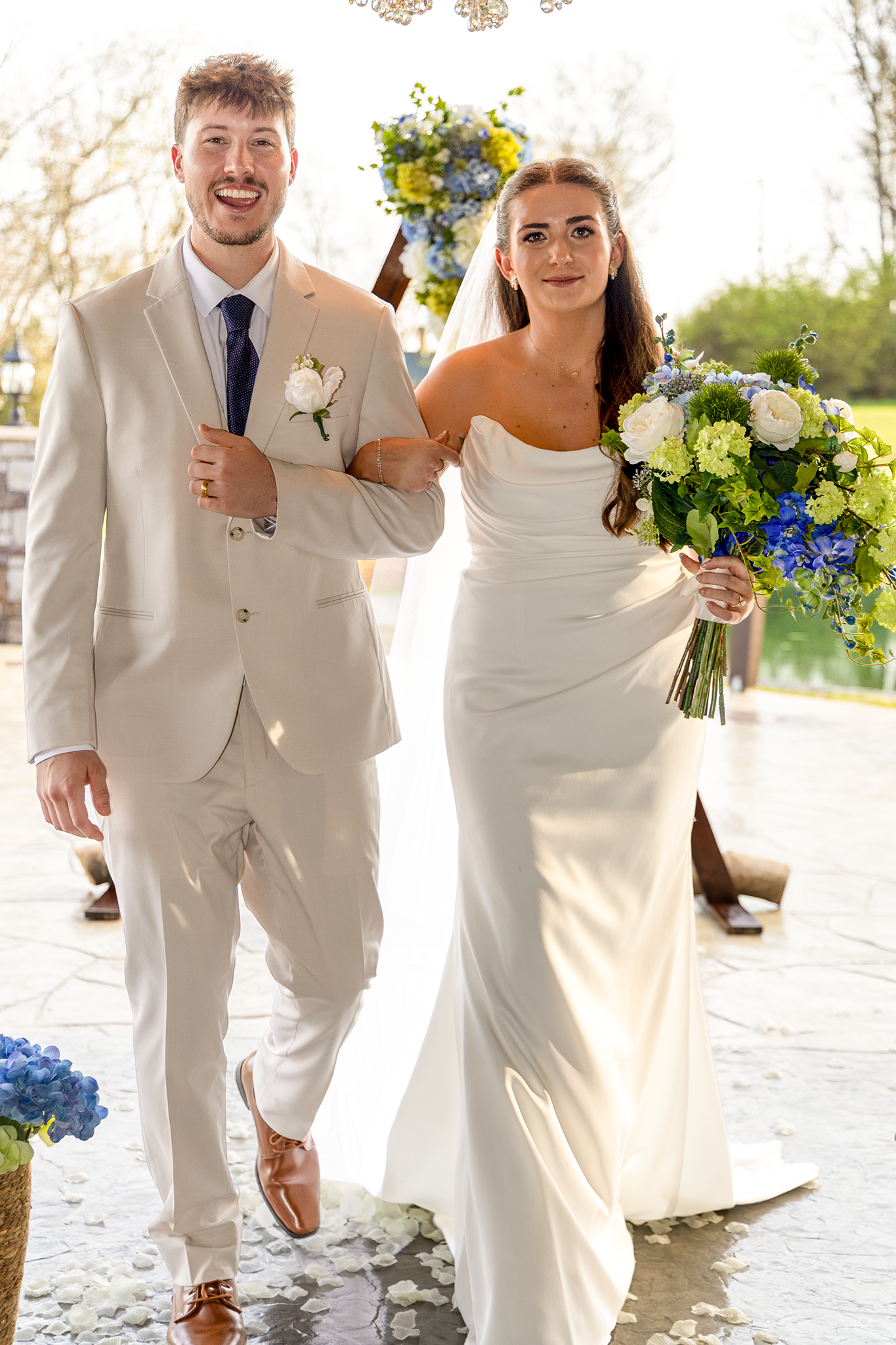 Beaming newlyweds walk arm in arm down the aisle after ceremony, bride carrying blue and green floral bouquet.