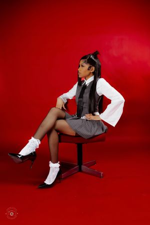 Young woman in school-inspired pinstripe outfit with white ruffle socks and heels posing on office chair against red backdrop.