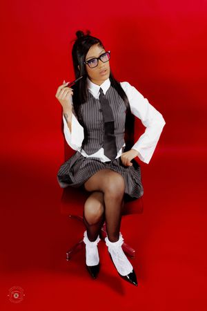 Woman in school uniform with glasses, vest, skirt, and stockings poses on chair against red background.