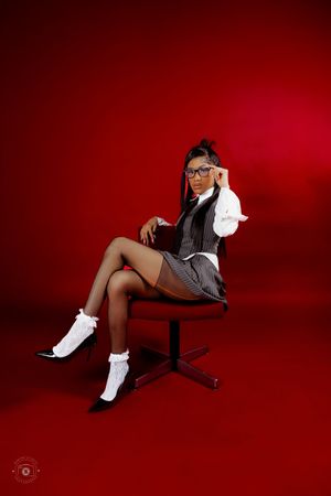 Woman in school-inspired outfit with glasses, white blouse, vest, plaid skirt, and ruffled socks seated on chair against red background.