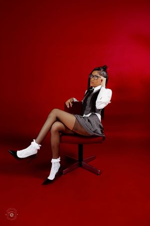 Woman in school uniform with glasses sits on chair against red background, wearing white socks and heels.