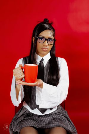 Young woman in school uniform with glasses holds a red mug, posing against a vibrant red background.