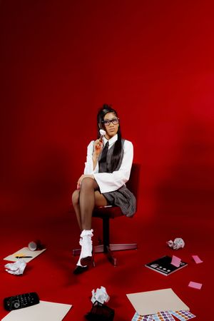 Young woman in school uniform sits on chair surrounded by scattered papers and makeup on red background.
