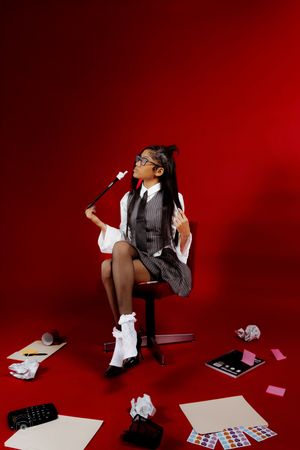 Young woman in business attire sits on chair surrounded by scattered papers and office supplies on red background.
