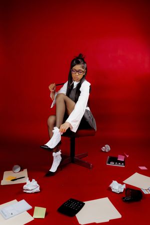 Young girl in glasses and office attire sits on chair surrounded by scattered papers and office supplies on red background.