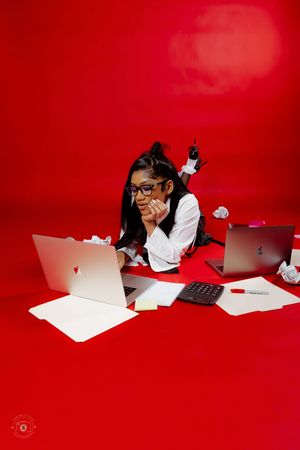 Young woman in glasses lying on red floor surrounded by laptops, crumpled paper, and office supplies.