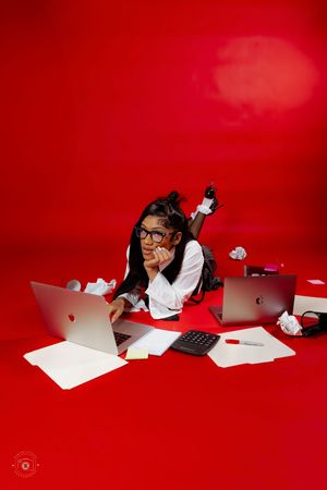 Young woman in glasses lying on red floor surrounded by laptops, papers, and a calculator in a busy workspace scene.