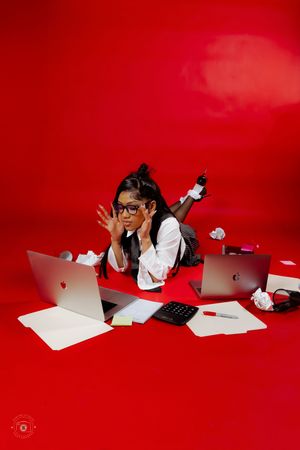 Woman in glasses lying on red floor surrounded by laptops, papers, and a calculator in a busy office concept photo.