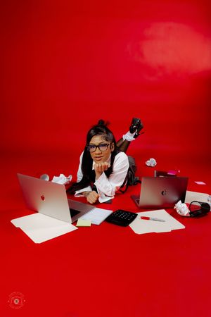 A young girl in glasses lies on a red floor surrounded by laptops, papers, and a calculator in a busy workspace scene.
