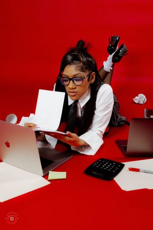 Young woman in office attire with glasses poses on red background with laptop, calculator, and papers.