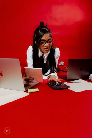 Young woman in glasses and vest working at red desk with laptops and calculator in professional setting.