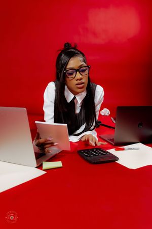 Young woman in glasses and white shirt works at red desk with laptops, tablet, and calculator in studio setting.