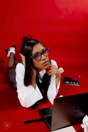 Young woman in glasses and business attire lying on red floor with laptop, looking thoughtful.