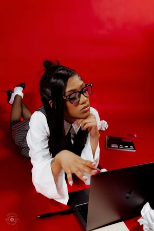 Young woman in glasses and business attire poses thoughtfully with a laptop against a bold red background.