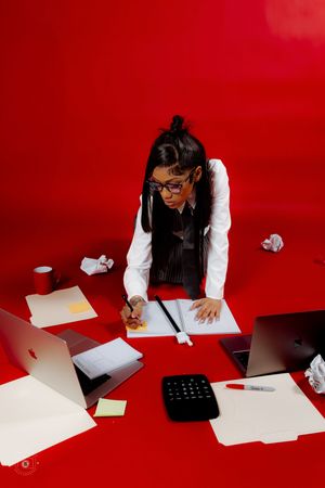 Woman in glasses leaning over red desk with laptops, papers, and calculator in a busy office workspace.