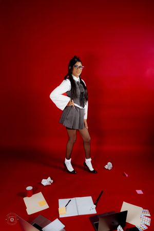 Young woman in school uniform with glasses poses confidently on red background surrounded by scattered papers and books.