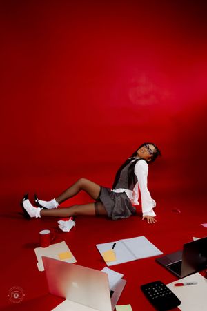 Woman in office attire lying on red floor surrounded by scattered papers, laptop, and calculator in creative photo shoot.