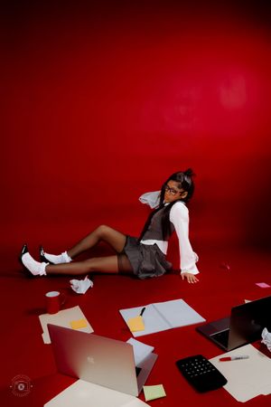 Young woman in school uniform sits on red floor surrounded by scattered papers, laptop, and office supplies in stylish photo shoot.