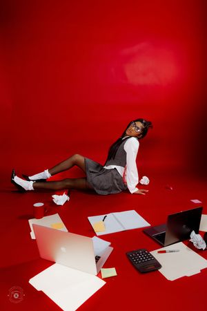 Woman in office attire relaxing on red floor surrounded by scattered papers, laptops, and office supplies.