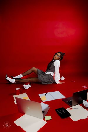 Woman in glasses sits on red floor surrounded by laptops, papers, and office supplies in a chaotic work scene.